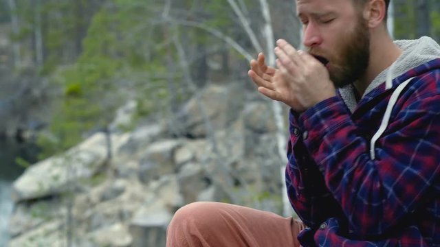 Beard man sitting on the rock near the lake and playing on the harmonica.