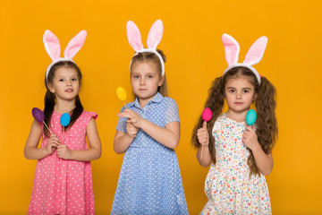 three little girls with Easter bunny ears holding colorful eggs on yellow background. Happy easter. cute children