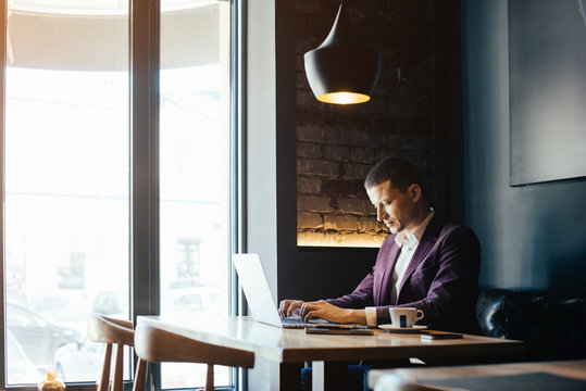 Young handsome man sitting in office with cup of coffee and working on project connected with modern cyber technologies. Businessman with notebook trying to keep deadline in digital marketing sphere.