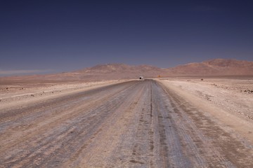 Fototapeta na wymiar Endless dirt road to infinity of salt flat plateau contrasting with blue cloudless sky. In the horizon isolated truck and yellow traffic sign showing left direction - Atacama desert , Chile