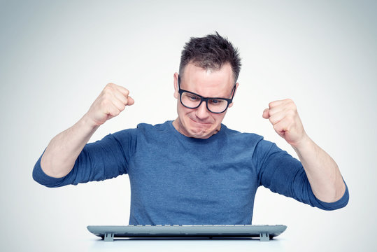 Upset man in glasses behind a keyboard in front of a computer, emotionally waving his fists at the keyboard. Failure concept
