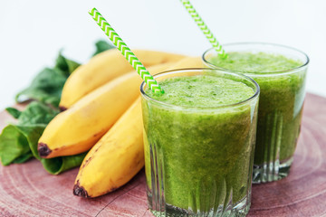 Two green smoothies in glasses with straws, banana and spinach, healthy ingredients on the wooden table