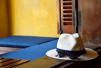 Sunglasses and beach hat on the window edge with yellow wall, wooden window frame and blue pillows in the background