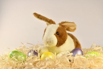 A plush bunny in front of a yellow egg in a nest with painted Easter eggs. Closer to the bottom in the center.