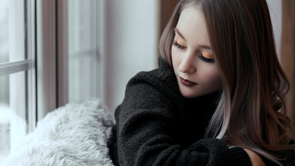 A beautiful young girl is sitting on the windowsill alone with her eyes closed. A woman in sweater sitting by the window