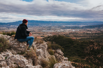 Young male sits on the rock and enjoys beautiful mountain landscape. Small city in background.