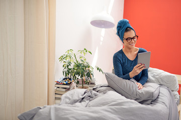 Gorgeous brunette with eyeglasses and towel on head using tablet while sitting on the bed in...