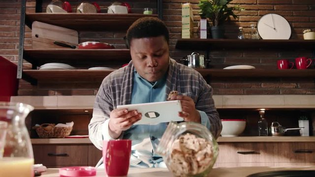 Serious african American man use digital tablet computer at kitchen eating homemade cookies relaxed american black home technology touchscreen wireless eating morning close up slow motion