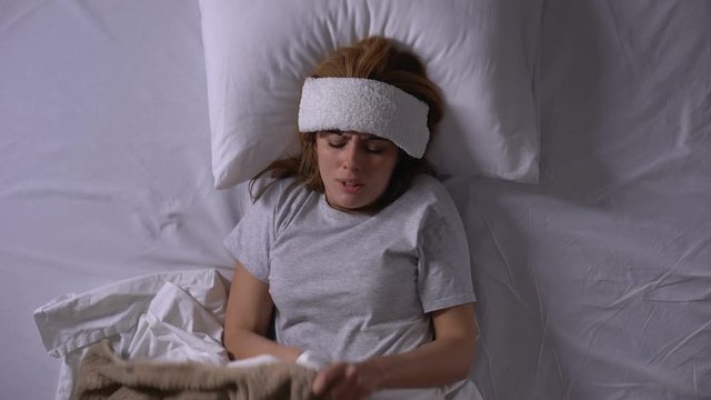 Young woman suffering from high fever, lying in bed with compress on forehead
