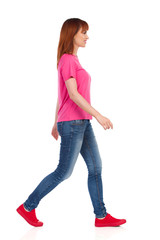 Fototapeta premium Side View Of Walking Young Woman In Pink Shirt, Jeans And Red Sneakers