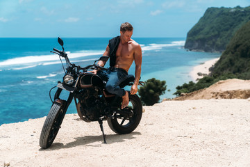 Fototapeta na wymiar Sexy athletic man with perfect naked body sitting on motorbike, ocean waves and beautiful mountains on background