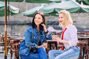 Portrait of two young women sitting together drinking coffee in summer street cafe. Girls photographing themselves with retro camera.