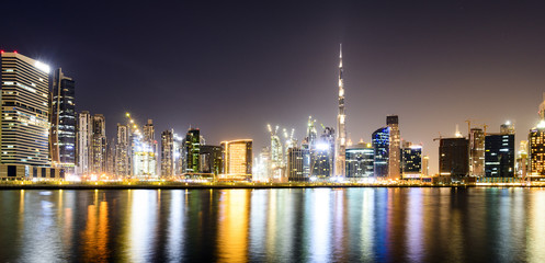 Fototapeta na wymiar Stunning view of the illuminated Dubai skyline during sunset with the magnificent Burj Khalifa and many other buildings and skyscrapers reflected on a silky smooth water flowing in the foreground.