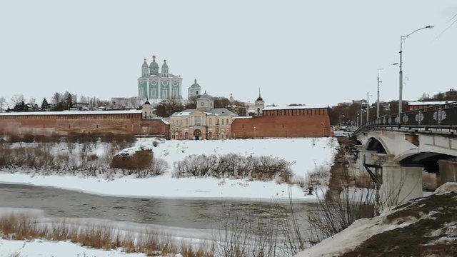 River flows in winter, on the bank is located palace, Orthodox church, fortress with red walls in russain city. There is snow on the street, an old bridge with black railings and lampposts passes acro