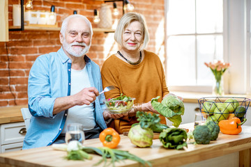 Cheerful senior couple eating salad standing together with healthy food on the kitchen at home. Concept of healthy nutrition in older age