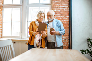 Beautiful senior couple using digital tablet standing together near the window at home
