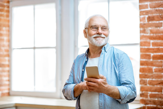 Cheerful senior grandfather using phone standing near the window at home