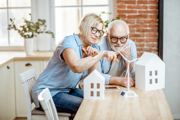 Senior couple dreaming about modern property and alternative energy, sitting with house models and...