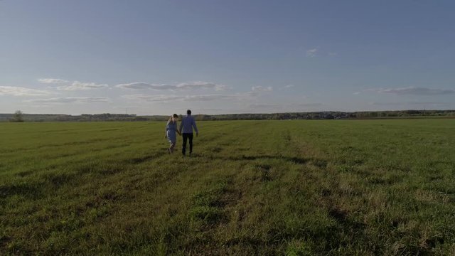 A man in a blue shirt and jeans holding the hand of a girl in a blue dress, they go on the field,aerial photography, the camera flies around a couple