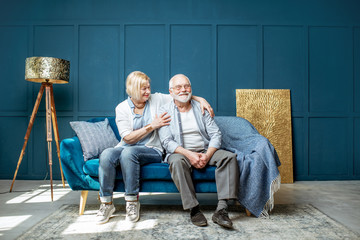 Lovely senior couple sitting together on the couch in the living room at home, wide interior view - obrazy, fototapety, plakaty