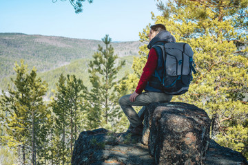 Naklejka premium Man hiker with a large backpack sitting on a rock cliff and looking at the sprawling green valley. Freedom in travel