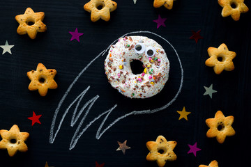 donut meteorite with googly eyes among the stars of cookies on a black chalkboard background, funny children's entertainment with food, space concept
