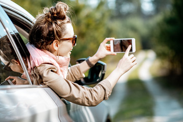 Young woman enjoying the trip, photographing out the window with phone on a picturesque road in the woods