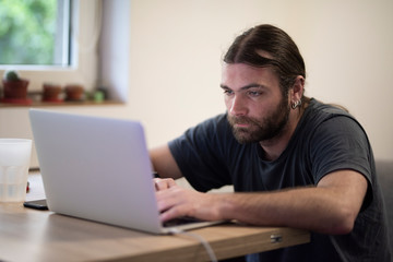 Young man with long hair using a laptop placed on a wooden desk while working from a cozy environment. Business require internet and portability most of the time