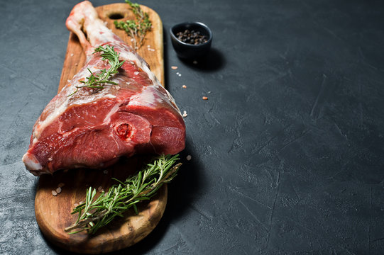 A raw leg of lamb on a wooden chopping Board. Rosemary, thyme, black pepper. Black background, side view, space for text