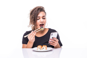 Smiling young woman using mobile phone eating sushi at the table isolated over white background,