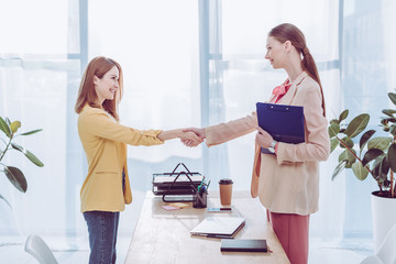 happy recruiter holding clipboard and shaking hands with woman in modern office