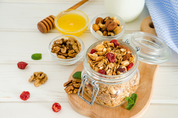 Multicereal homemade organic granola with a mixture of nuts, dried cherries, honey, cinnamon  and yogurt in a glass jar on a white wooden background.