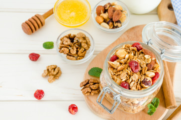 Multicereal homemade organic granola with a mixture of nuts, dried cherries, honey, cinnamon  and yogurt in a glass jar on a white wooden background.