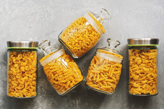 Flat lay a variety of uncooked pasta in glass jars on a gray background. Top view