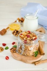 Multicereal homemade organic granola with a mixture of nuts, dried cherries, honey, cinnamon  and yogurt in a glass jar on a white wooden background.