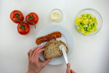 A woman is making a BLT sandwich on whole grain bread with bacon, lettuce, mayonaise and tomatoes on a white background.