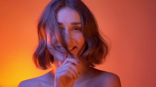 Closeup shoot of young attractive caucasian female with naked shoulders and playing with her hair looking at camera with neon red background