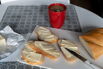 Red cup of coffee with milk and slices of bread with butter ( sandwich) and knife on the white board on the table in kitchen in the morning. Tasty breakfast. 