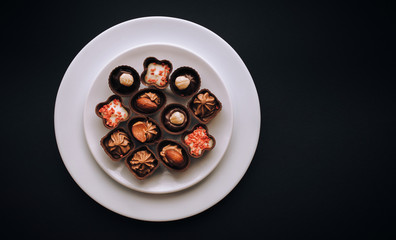 Many beautiful chocolates of different shapes and fillings lie in a white plate against the black background paper.