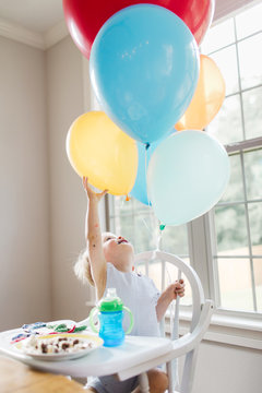 toddler reaches up to touch birthday balloons attached to his high chair