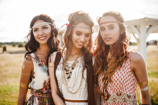 Three cute beautiful hippie girl in the setting sun, outdoors, the best of friends smiling and having fun, makeup, long hair, feathers in their hair, bracelets, flash tattoo, indie, Bohemia boho style