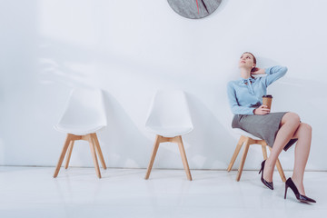 attractive woman holding paper cup and looking at clock while sitting on chair