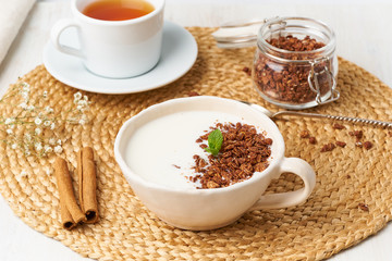 Yogurt with chocolate granola in cup, breakfast with tea on beige background, side view.