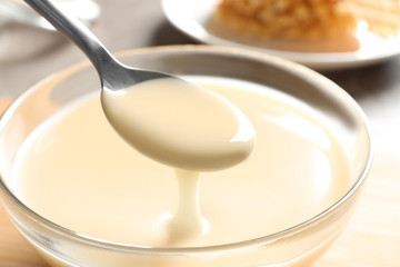Spoon of pouring condensed milk over bowl on table, closeup. Dairy products