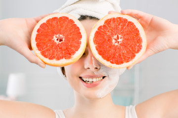 Young charming girl biting lips, has white fasial mask, holds two slices of grapefruit, has white towel on head. Natural cosmetics, skincare, wellness, facial treatment, cosmetology concept.
