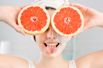 Young cheerful european woman with white facial mask and white towel on head shows tongue and holds big grapefruits, covering her eyes. Healthy diet, skincare, facial treatment, cosmetology concept.