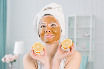 Beauty Skin Care Concept. Attractive Caucasian woman in white towel with brown facial mask on face holds citrus fruit on her hand on light background. Spa procedures and cream mask on skin