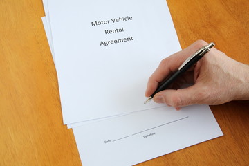 Motor vehicle rental agreement and hand with pen about to sign it on wooden table