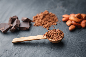 Cacao powder in a wooden spoon with cacao beans and chocolate on a dark background