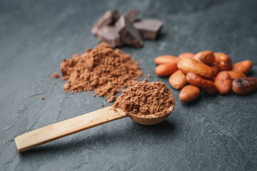 Cacao powder in a wooden spoon with cacao beans and chocolate on a dark background